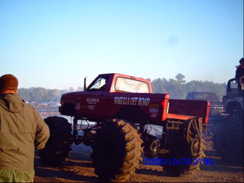 1987 Suzuki Samurai - Monster truck event with a red truck and other vehicles in the background under a clear sky.