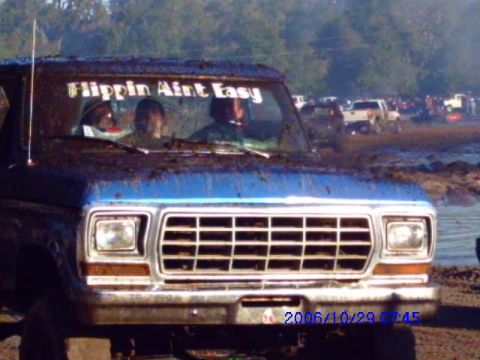 1987 Suzuki Samurai - A blue pickup truck is covered in mud, with people inside and text on the windshield.