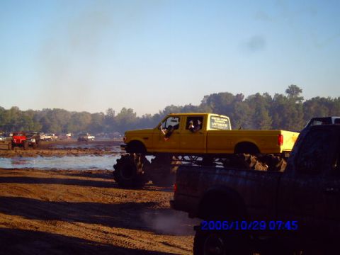 1987 Suzuki Samurai - A yellow monster truck navigates a muddy terrain with other vehicles visible in the background.