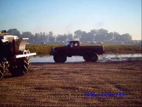 1987 Suzuki Samurai - Two modified trucks are seen in a muddy field during a daytime event. The truck on the right is a dark red pickup.