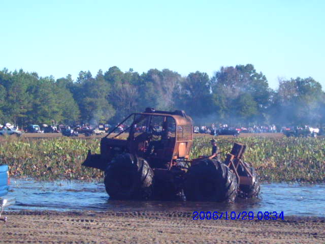 Custom mud bogger with large tires in water, trees and spectators in background
