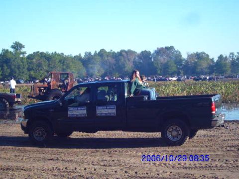 1987 Suzuki Samurai - A black pickup truck is parked on a dirt area with a woman sitting in the bed. Multiple vehicles are visible in the background.