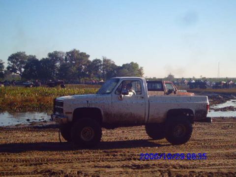 1987 Suzuki Samurai - A white pickup truck is stuck in the mud with other vehicles visible in the distance.
