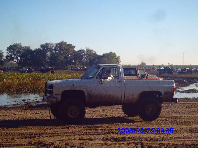Pickup truck with large tires in a muddy field, other vehicles and trees in background