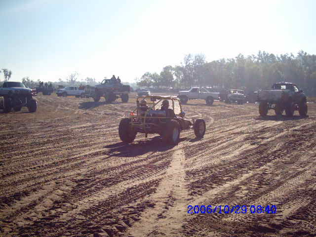 Several off-road vehicles, including a dune buggy and lifted trucks, are parked and driving in a sandy, tire-tracked area under a clear sky
