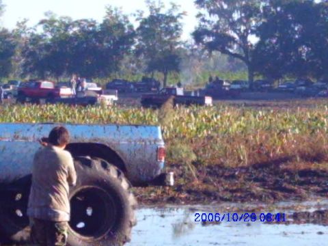 1987 Suzuki Samurai - A muddy blue pickup truck with oversized tires sits in a field, with several other vehicles visible in the distance.