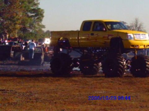 1987 Suzuki Samurai - A yellow monster truck with flame decals is shown in a muddy outdoor area with spectators in the background.