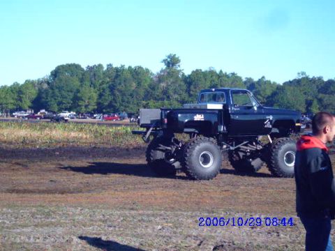 1987 Suzuki Samurai - A modified black truck with oversized tires sits in a muddy area under a clear blue sky.