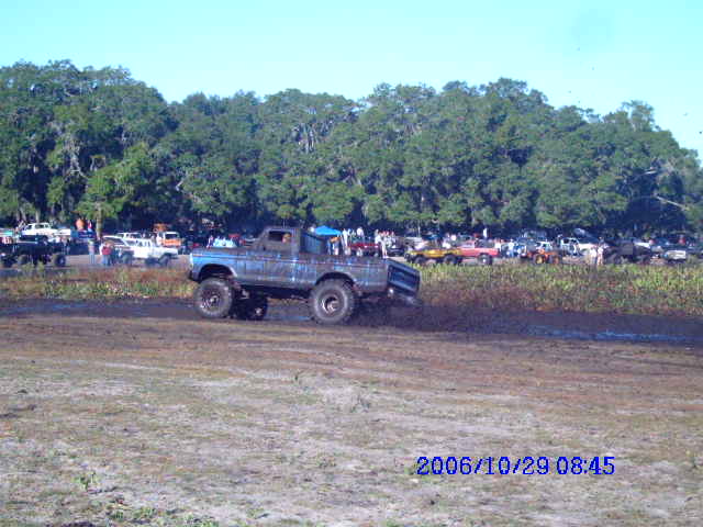 Big blue lifted truck driving through mud, other vehicles and trees in background