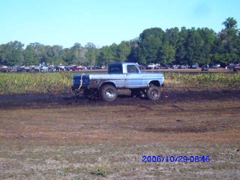 1987 Suzuki Samurai - A blue pickup truck navigates a muddy track, with spectators and other vehicles visible in the distance.