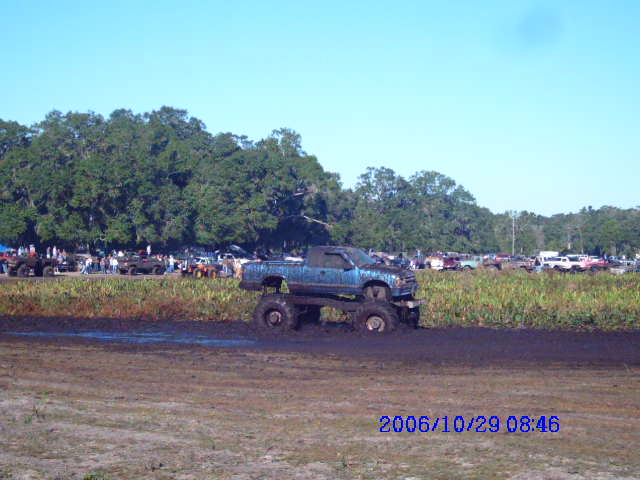 Blue pickup truck with large tires stuck in mud, trees and other vehicles in background
