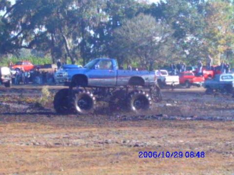 1987 Suzuki Samurai - A blue monster truck is shown in a muddy area with other vehicles and spectators present.