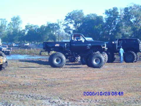 1987 Suzuki Samurai - Two large trucks are parked on a muddy field with trees in the background.