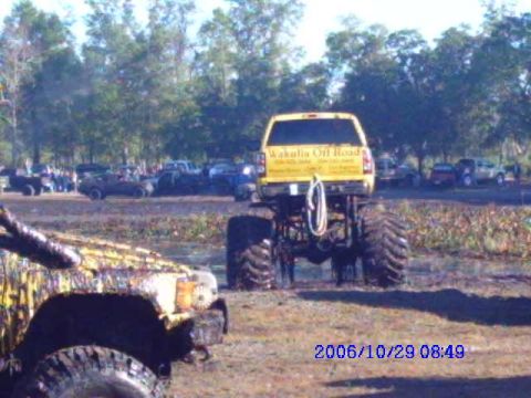 1987 Suzuki Samurai - A yellow pickup truck modified with oversized tires is seen in a muddy outdoor area with other vehicles in the background.