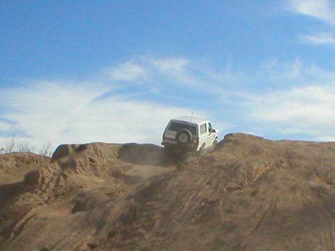 1993 Ford Bronco - A white SUV navigates a challenging dirt terrain, kicking up dust as it ascends the hill.