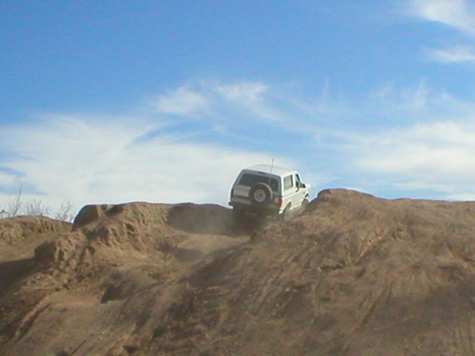 White Ford Bronco cresting a steep dirt berm in the desert, rear view kicking up dust