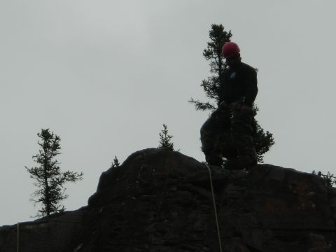 2003 Jeep TJ - A person wearing a red helmet and camouflage pants rappels down a rocky outcrop under an overcast sky.