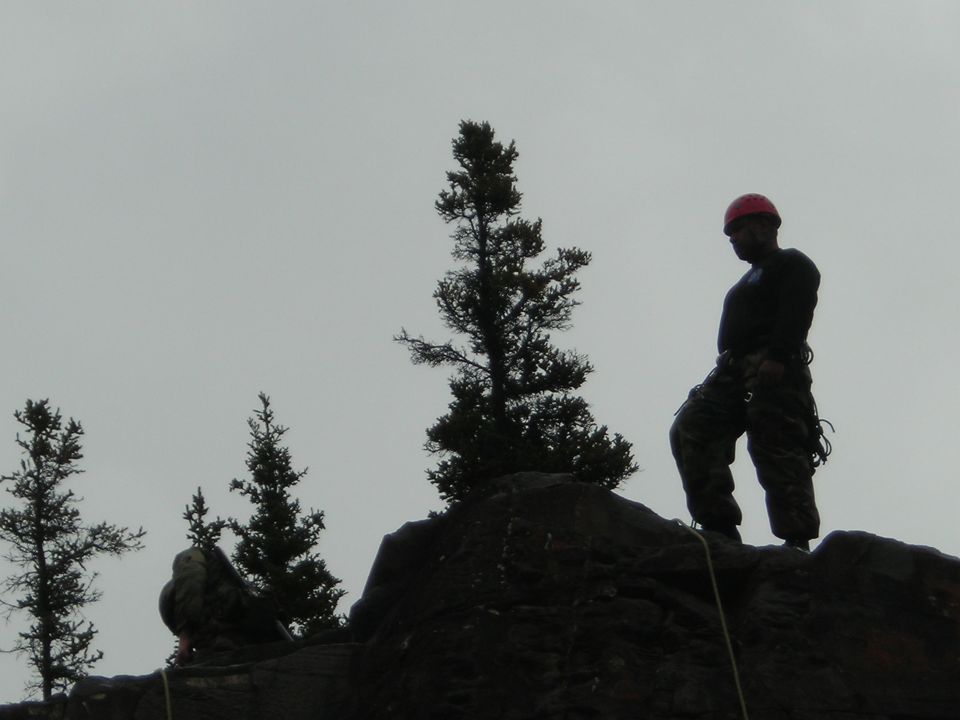 Two people in climbing gear on a rocky outcrop with trees under a cloudy sky