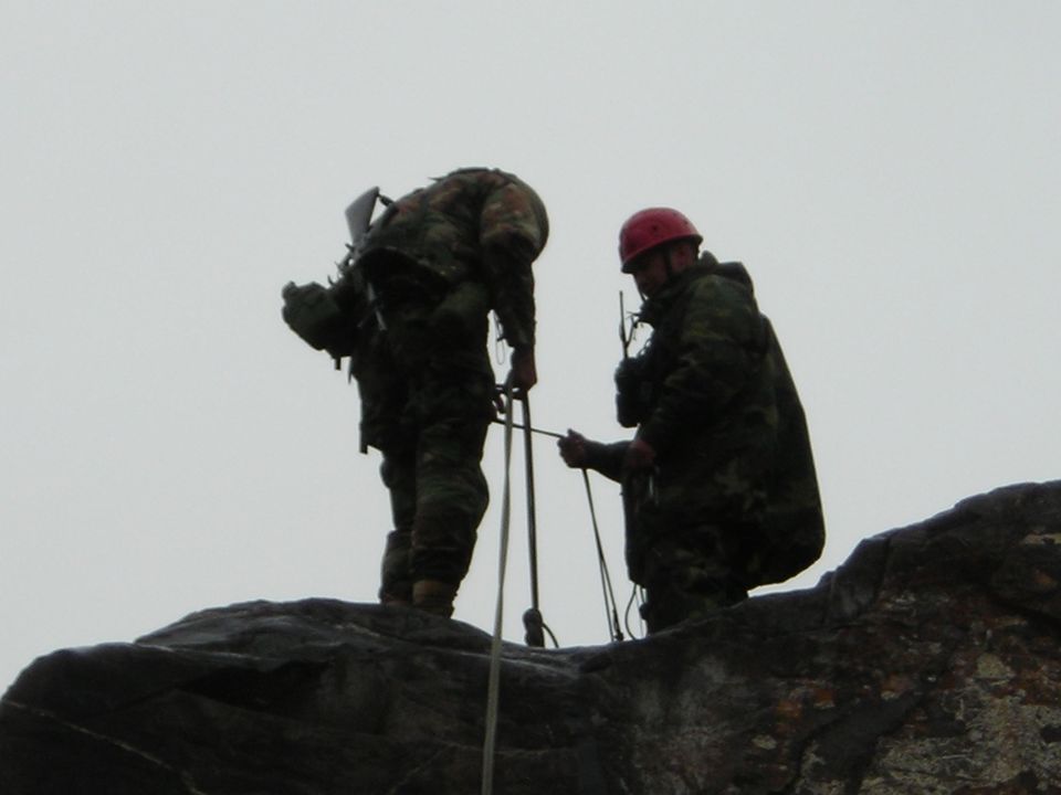 Two people in camouflage gear and one wearing a red helmet are rappelling down a rocky cliff face