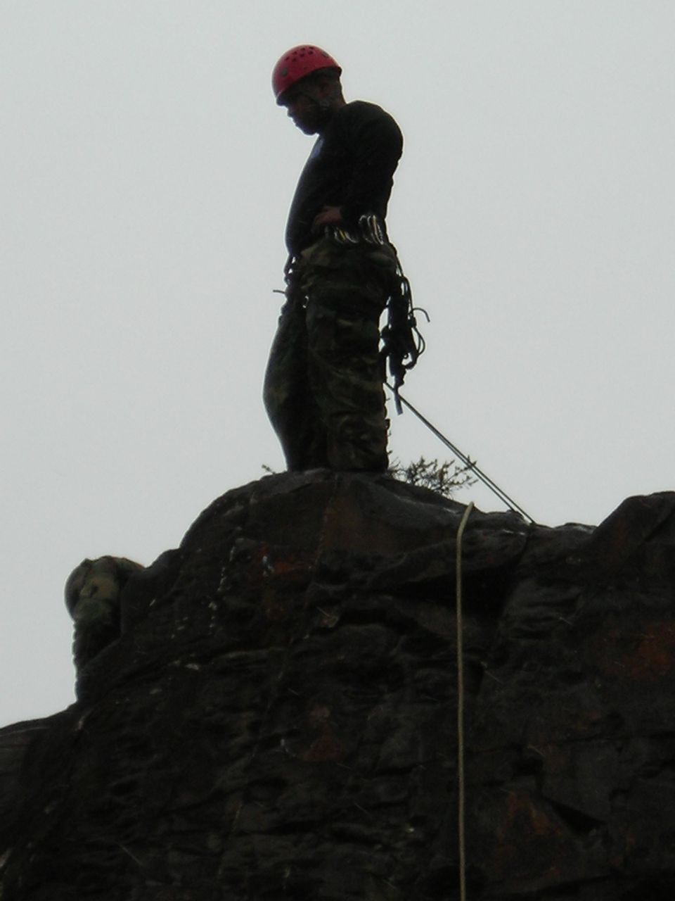 Climber in red helmet and camouflage pants stands on a dark rock formation against a gray sky