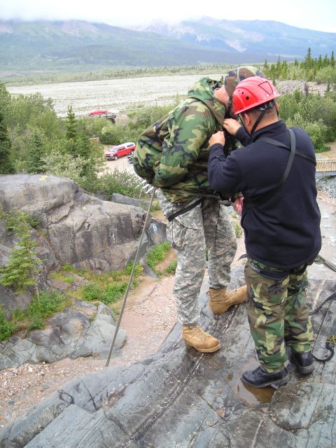 2003 Jeep TJ - Two individuals are geared up for rappelling on a rocky outcrop, with vehicles in the distance.