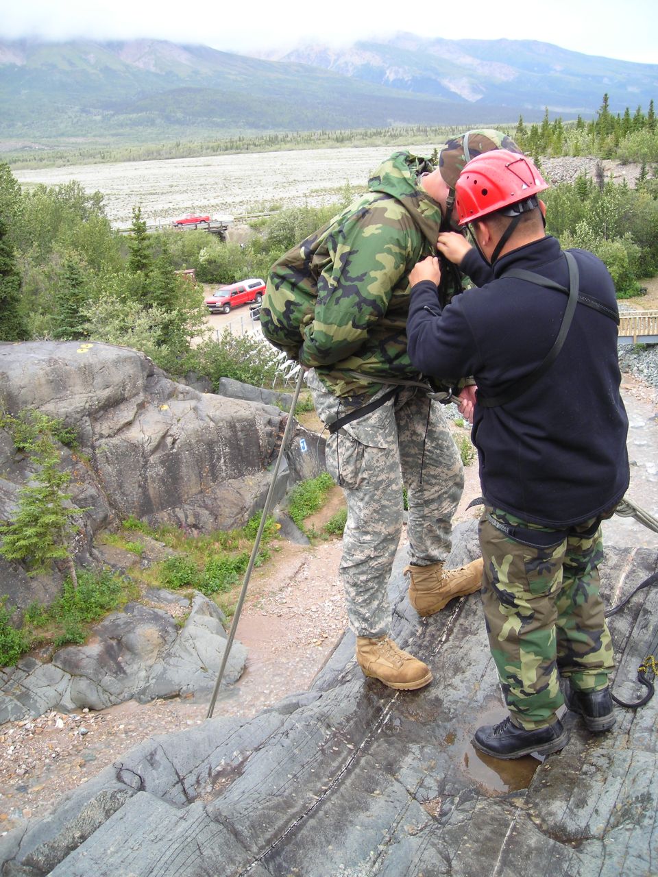 Two people in camouflage gear are preparing to rappel down a rocky cliff. A red SUV and a red truck are visible in the background