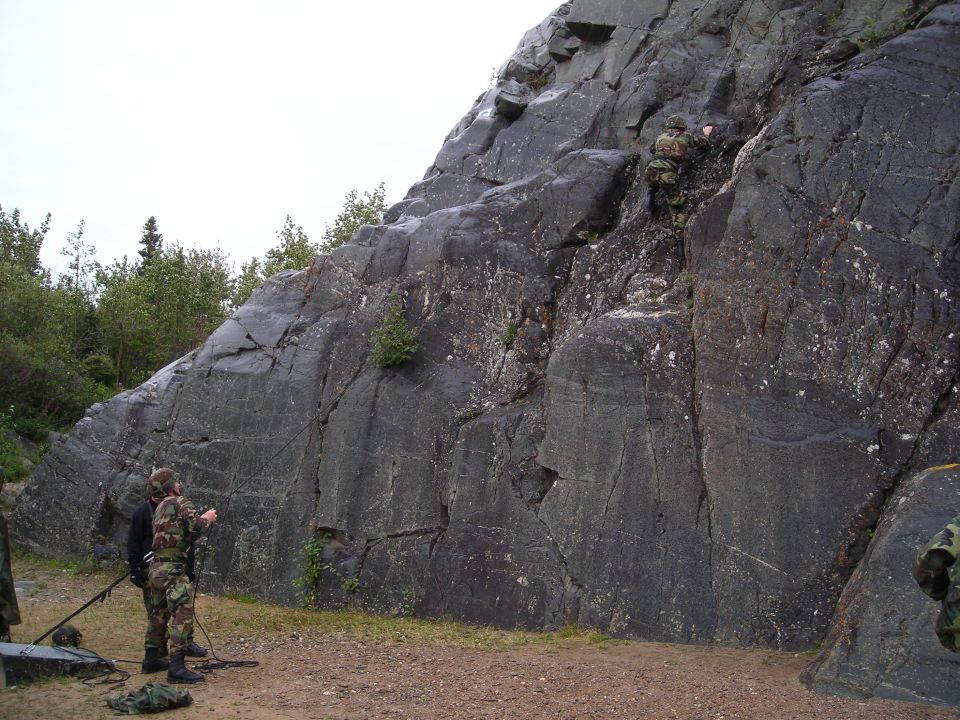 Rock face with two people in camouflage gear standing at the bottom and one person climbing