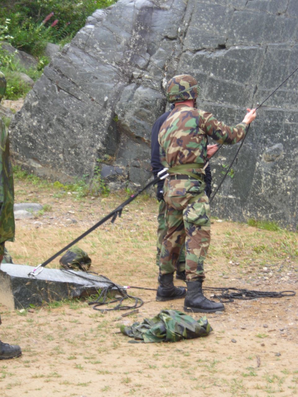 Military personnel in camouflage gear prepare ropes for rappelling next to a large rock face