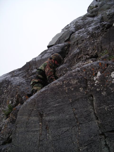 2003 Jeep TJ - A person in camouflage gear ascends a steep, textured rock formation on a cloudy day.