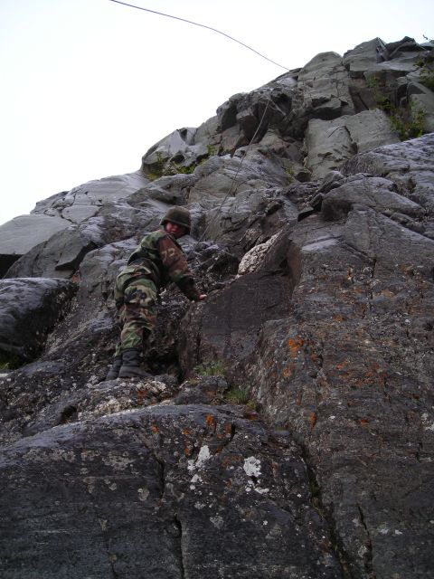 2003 Jeep TJ - A person in camouflage gear ascends a steep, rocky incline, with a rope visible against the overcast sky.