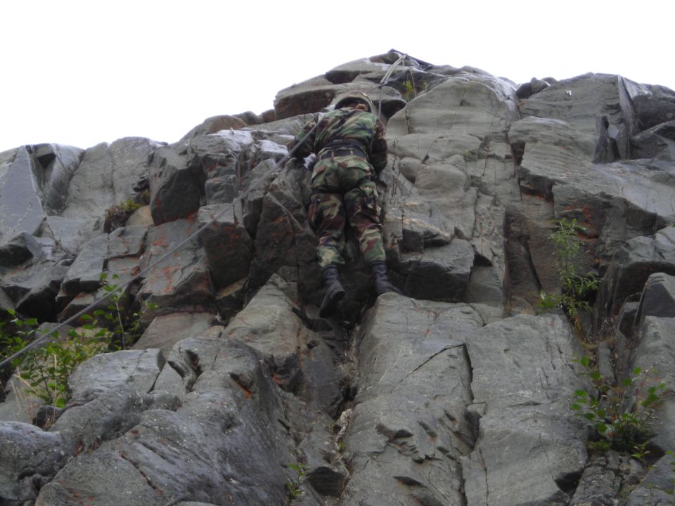 Soldier in camouflage climbing a steep, gray rock face with ropes