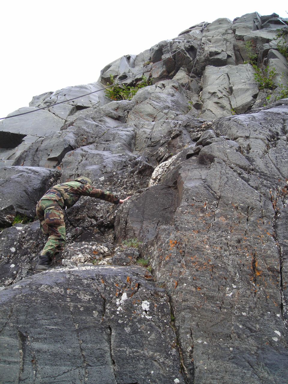 Person in camouflage clothing climbing a steep, grey, rocky cliff face with some sparse green vegetation