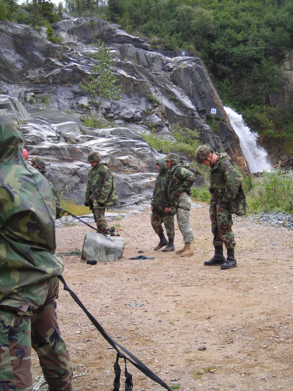 Soldiers in camouflage stand near a rocky cliff with a waterfall. One soldier holds a rope