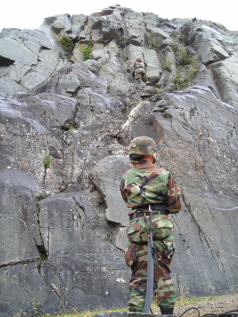Soldiers in camouflage gear ascend a steep, rocky cliff face with ropes