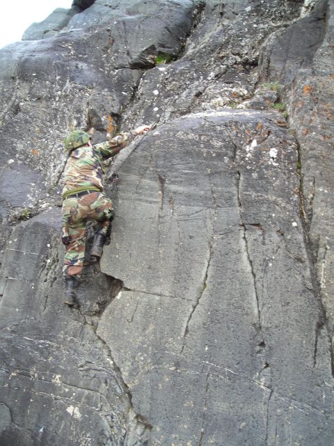 2003 Jeep TJ - A person in camouflage gear ascends a steep rock formation, using handholds and footholds.