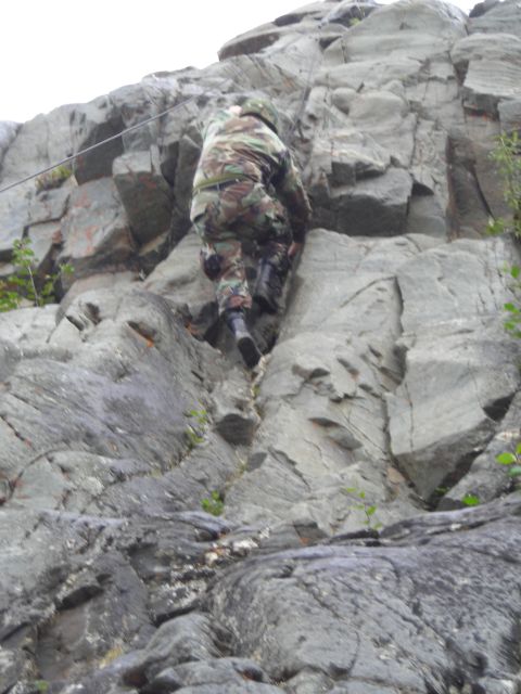 2003 Jeep TJ - A person in camouflage gear ascends a challenging rock face, utilizing a safety cable for support.