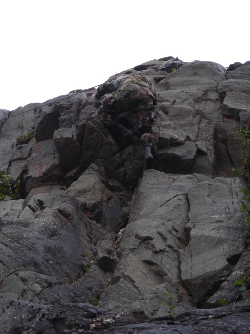 person in camouflage climbing a steep, rocky cliff face with a rope