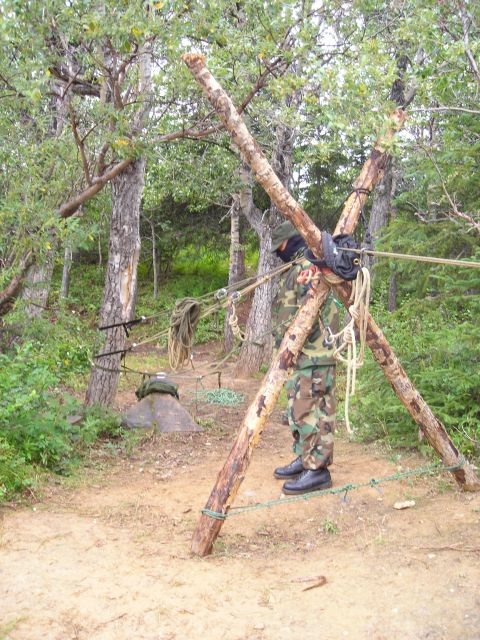 2003 Jeep TJ - A person in camouflage attire is positioned near a makeshift wooden tripod structure with ropes, set up in a forest environment.