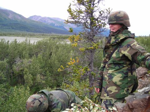 2003 Jeep TJ - Two individuals in military camouflage uniforms are positioned in a natural, wooded environment with a mountainous backdrop.