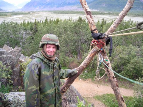 2003 Jeep TJ - A person in camouflage gear stands outdoors near a wooden structure with ropes, overlooking a valley.