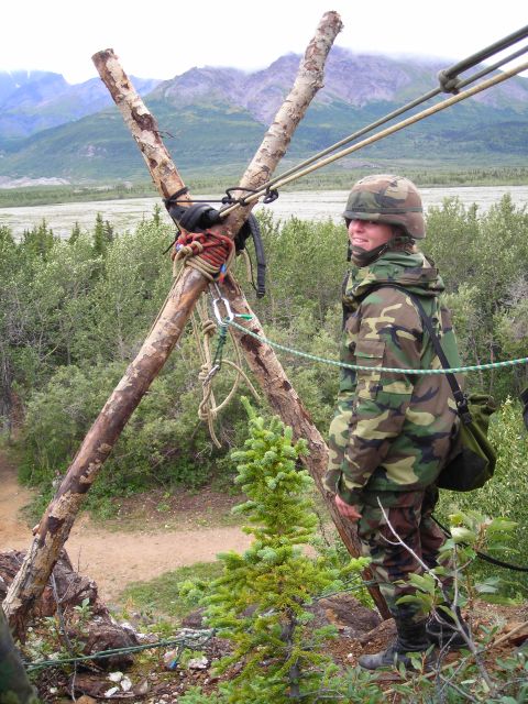 2003 Jeep TJ - A person in military-style camouflage stands on a rocky outcrop next to a wooden structure with ropes, with a scenic mountain landscape in…