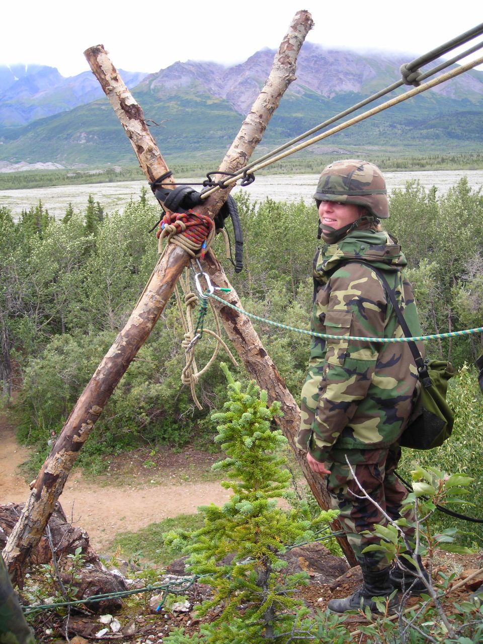 Person in camouflage gear stands near a wooden tripod with ropes, overlooking a valley and mountains