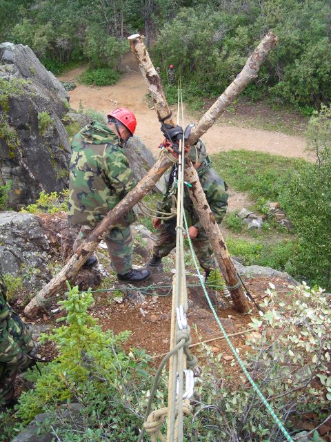 2003 Jeep TJ - Two individuals in camouflage gear are assembling a tripod structure using logs and ropes on a rocky, outdoor terrain.