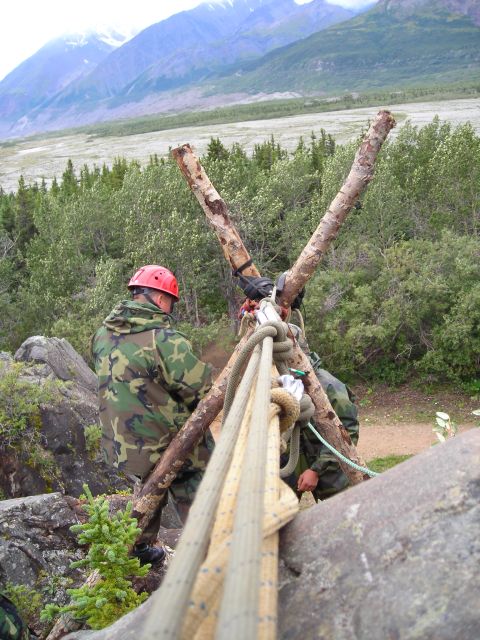 2003 Jeep TJ - Individuals in camouflage gear are engaged in an activity involving ropes and logs, set against a backdrop of mountains and trees.
