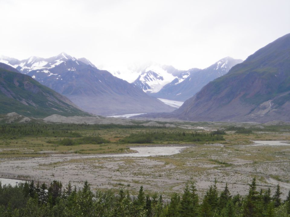 Wide mountain valley with a glacier in the distance, riverbed in foreground, trees at bottom