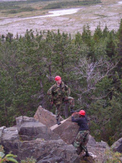 2003 Jeep TJ - Two individuals in camouflage gear and red helmets are positioned on a rocky outcrop overlooking a river valley.