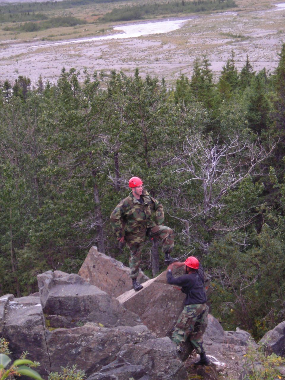 Two people in camouflage uniforms and red helmets stand on large rocks with a wooded landscape and river in the background