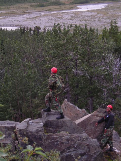 2003 Jeep TJ - Two individuals in camouflage gear and red helmets are positioned on a rocky outcrop, surveying a wide river valley below.