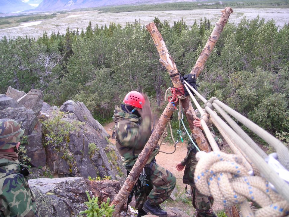 People in camouflage and red helmet setting up ropes on a wooden tripod structure on a rocky outcrop