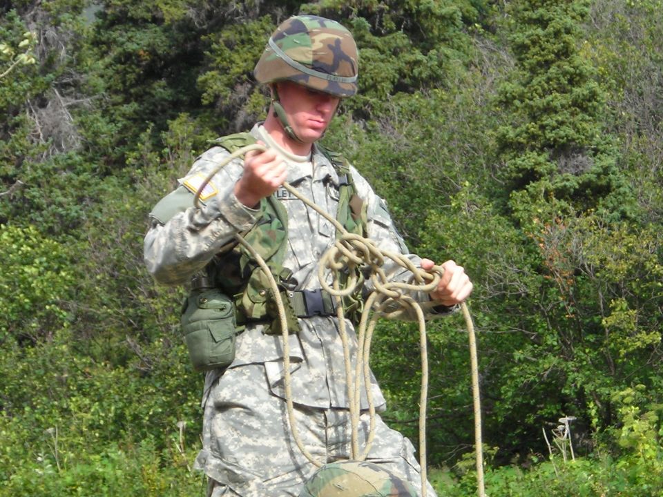Soldier in camouflage uniform and helmet tangles a thick rope in his hands, with trees in the background